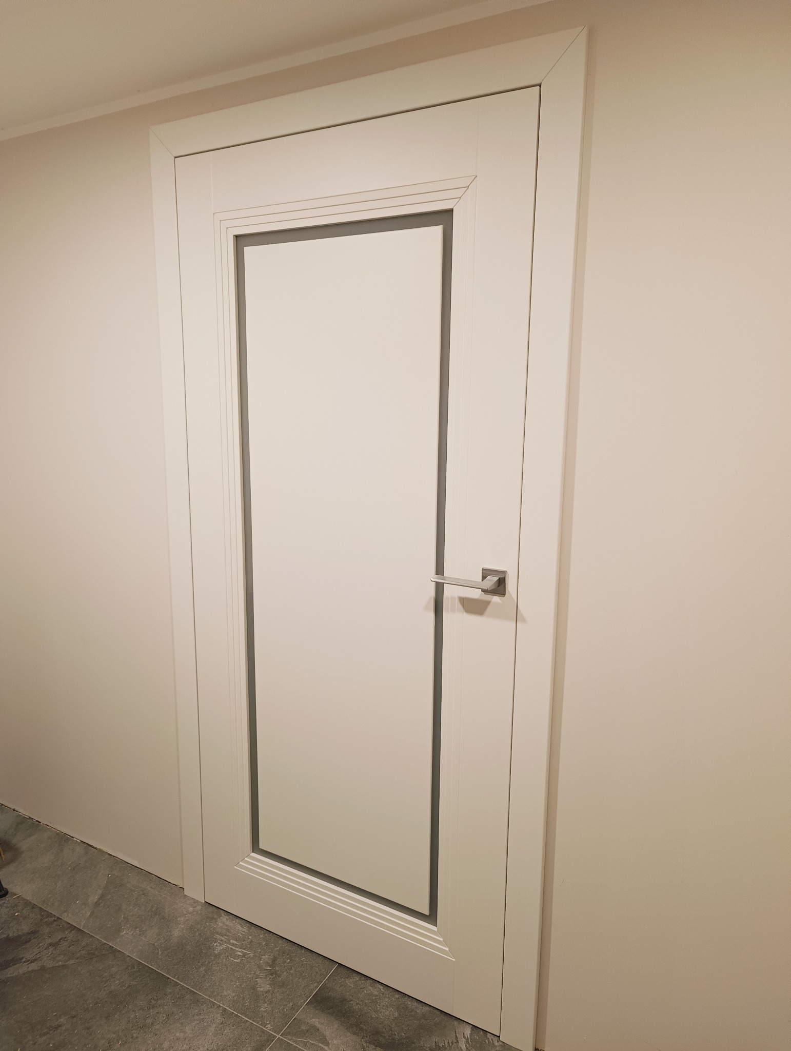 Zamknięte, białe drzwi wewnętrzne z szarą obwódką i srebrną klamką, zamontowane w jasnej ścianie. Widoczny fragment szarej podłogi. Minimalistyczny design.