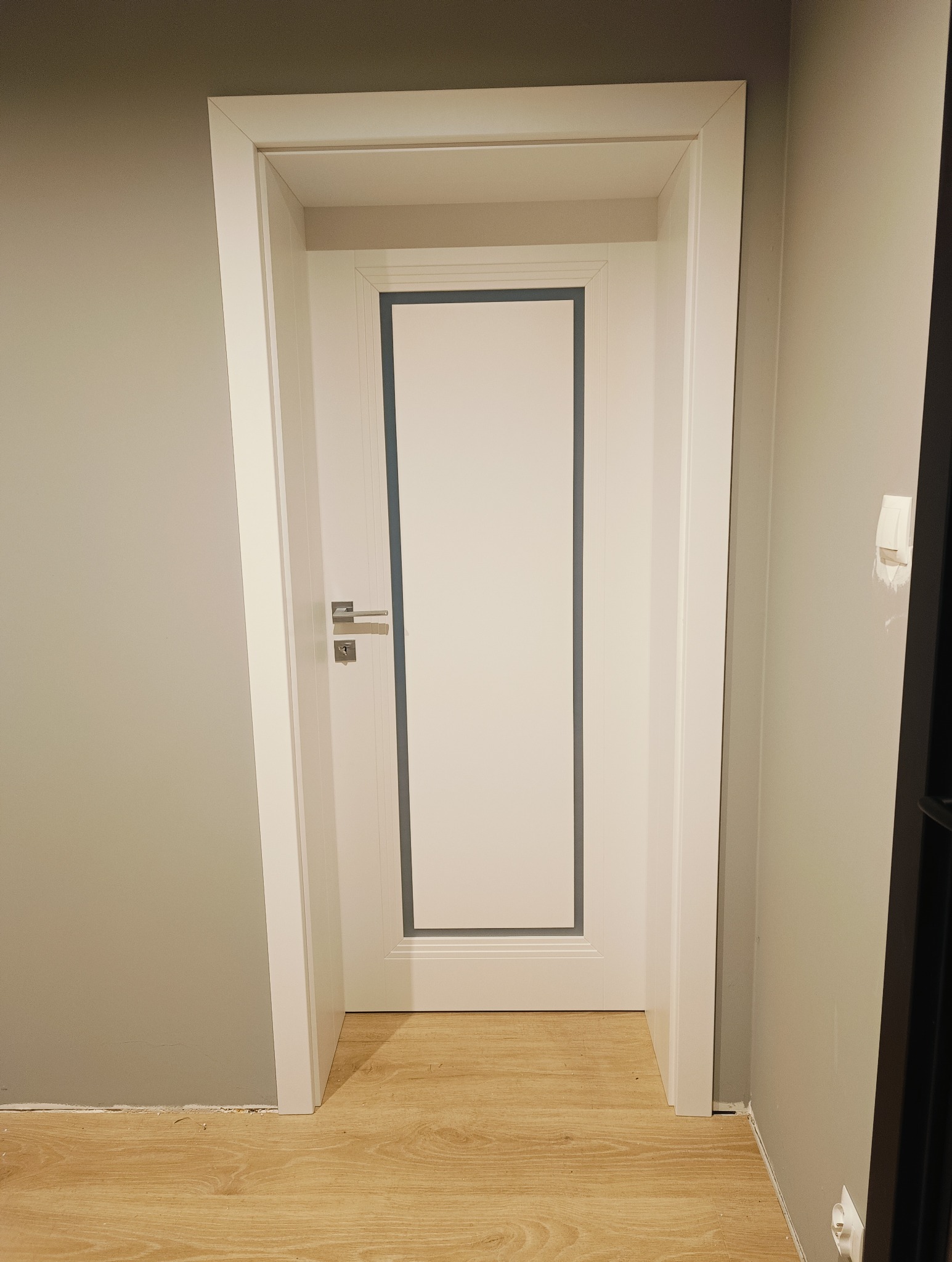 Białe drzwi wewnętrzne z szarym obramowaniem, zamontowane w szarej ścianie na drewnianej podłodze. Widoczna klamka i zamek. Minimalistyczny design.