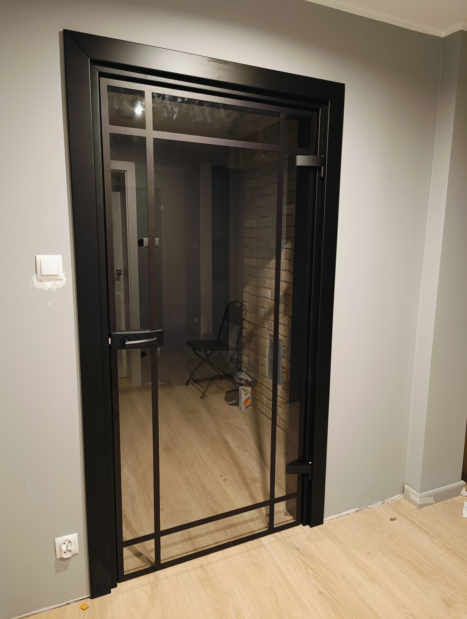 Nowoczesne drzwi wewnętrzne z czarną ramą i szklanymi panelami, widok na wnętrze z ceglaną ścianą i krzesłem. Minimalistyczny design, jasne drewniane podłogi.