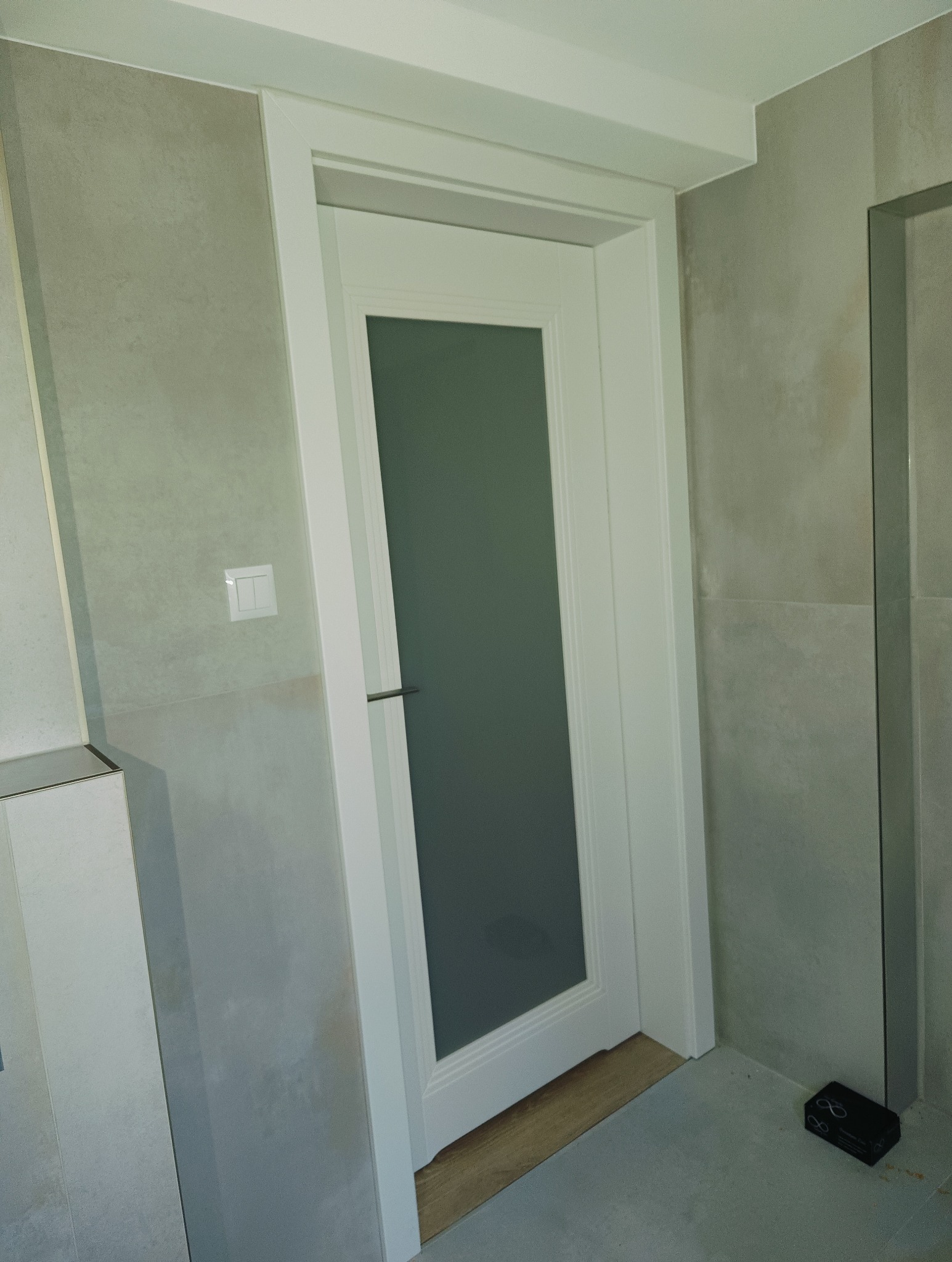 Białe drzwi wewnętrzne z matową szybą, zamontowane w łazience z szarymi płytkami. Widoczny fragment podłogi i ściany z włącznikiem. Minimalistyczny design.