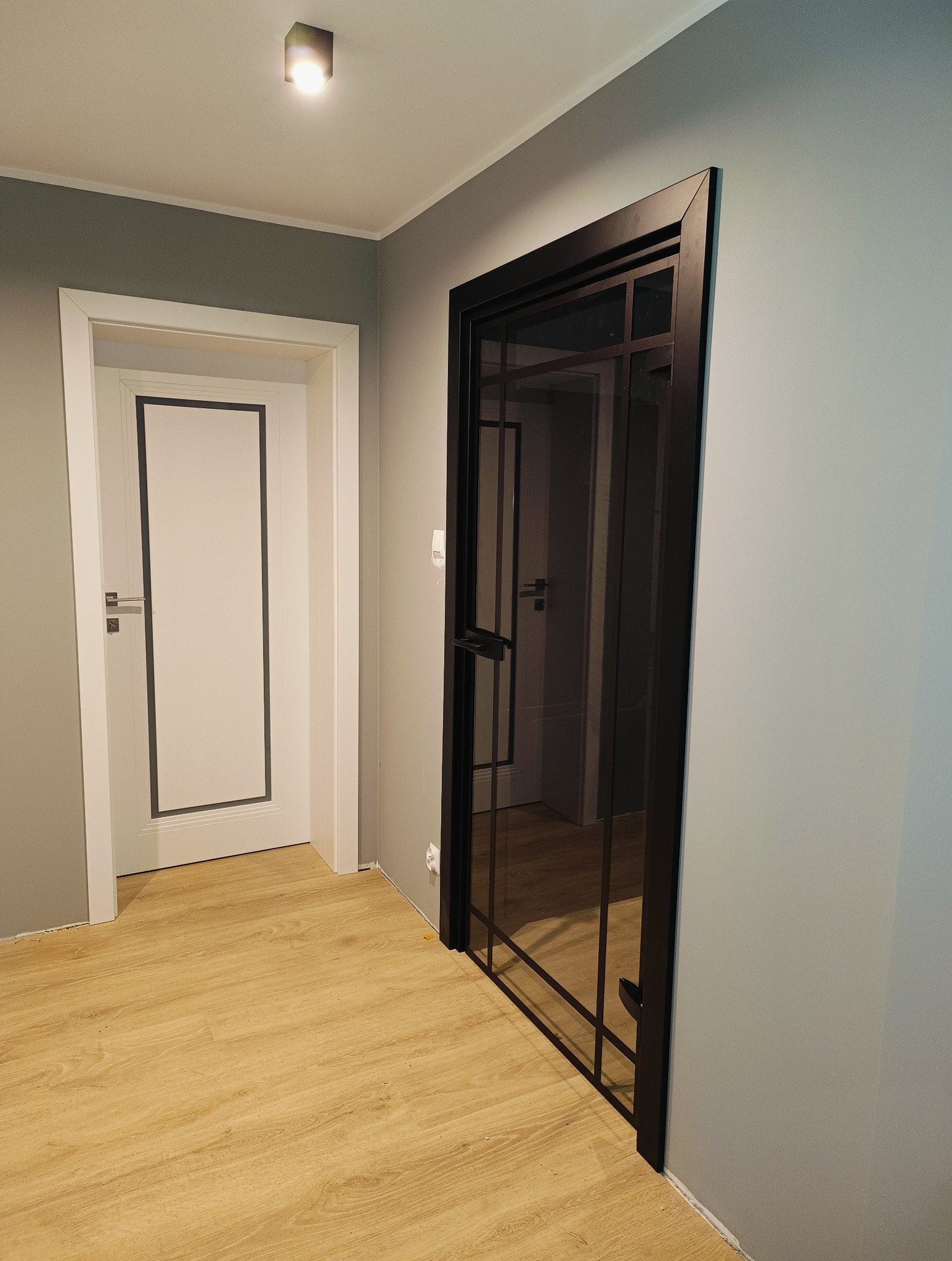 Nowoczesne, czarne drzwi wewnętrzne z przeszkleniami w stylu loft, widoczne na tle jasnych ścian i drewnianej podłogi. W tle białe drzwi z ciemną obwódką.