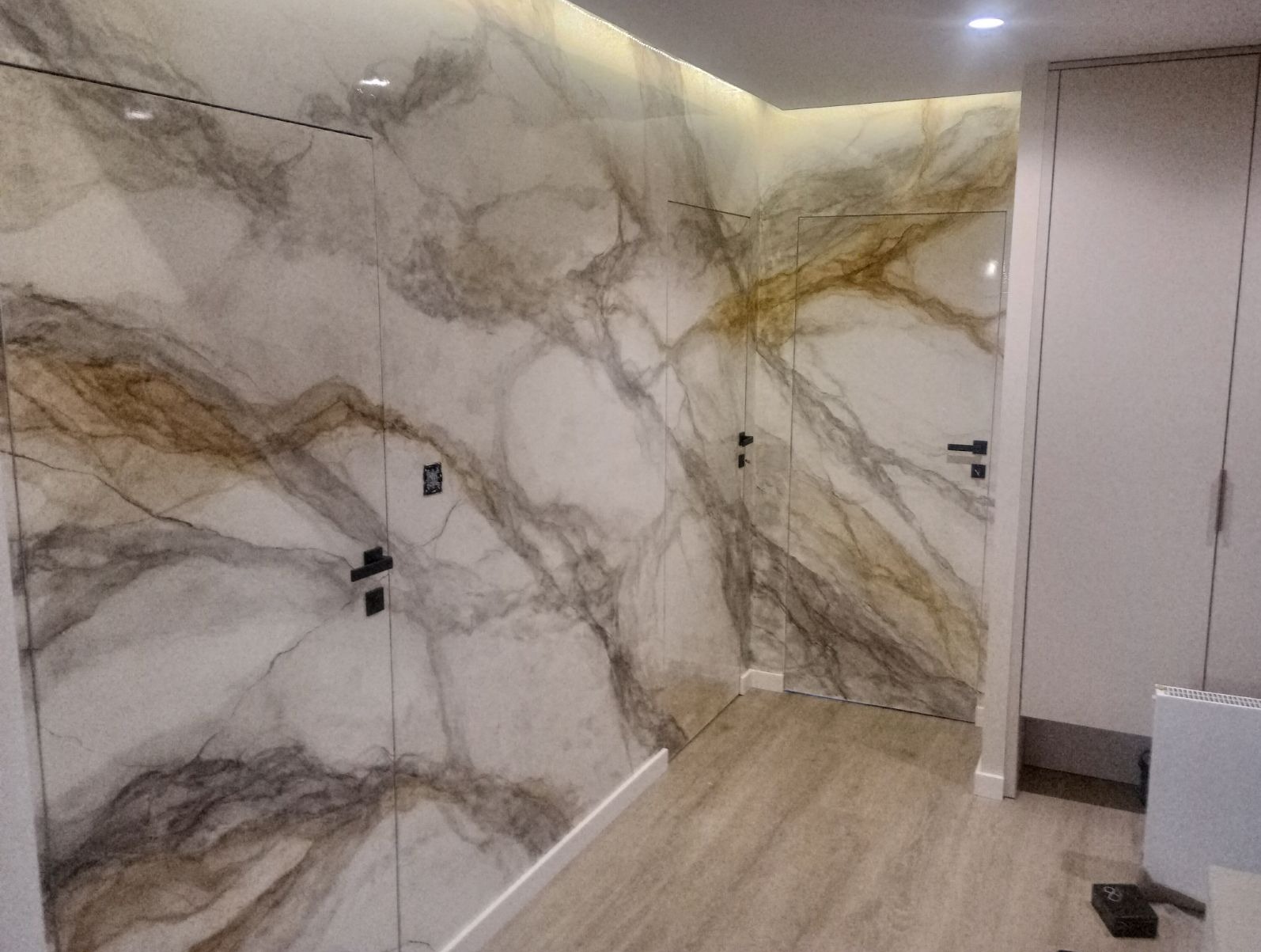 Dwa ukryte skrzydła drzwiowe z czarnymi klamkami, zlicowane ze ścianą o wzorze marmuru, w jasnym wnętrzu. Minimalistyczny design, nowoczesne wykończenie.