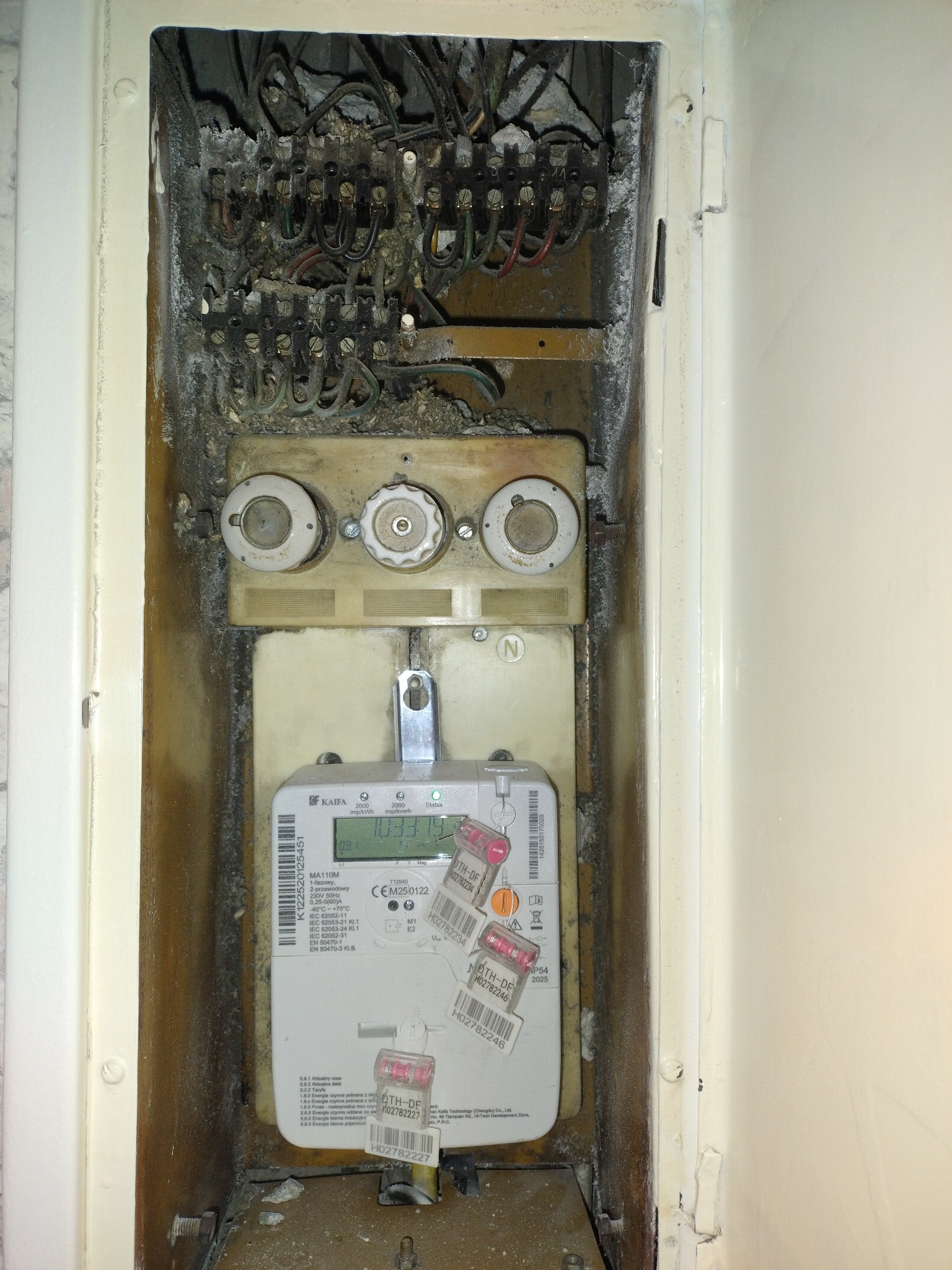 Otwarta skrzynka elektryczna z licznikiem, widoczne stare bezpieczniki topikowe i okablowanie. Ślady korozji i zabrudzeń wewnątrz obudowy.