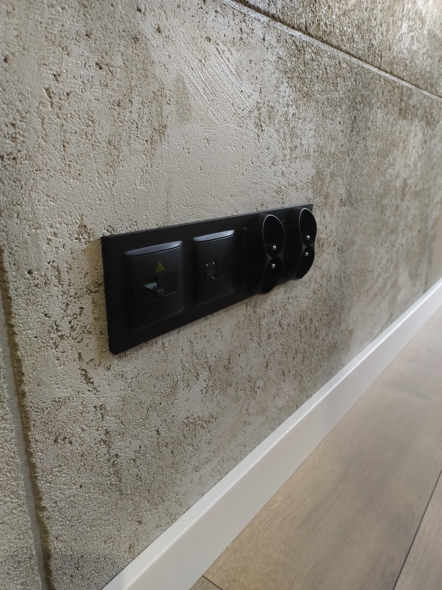 Czarny panel z gniazdkami i włącznikiem na teksturowanej ścianie, widok z boku, listwa przypodłogowa w kolorze białym, jasna podłoga.