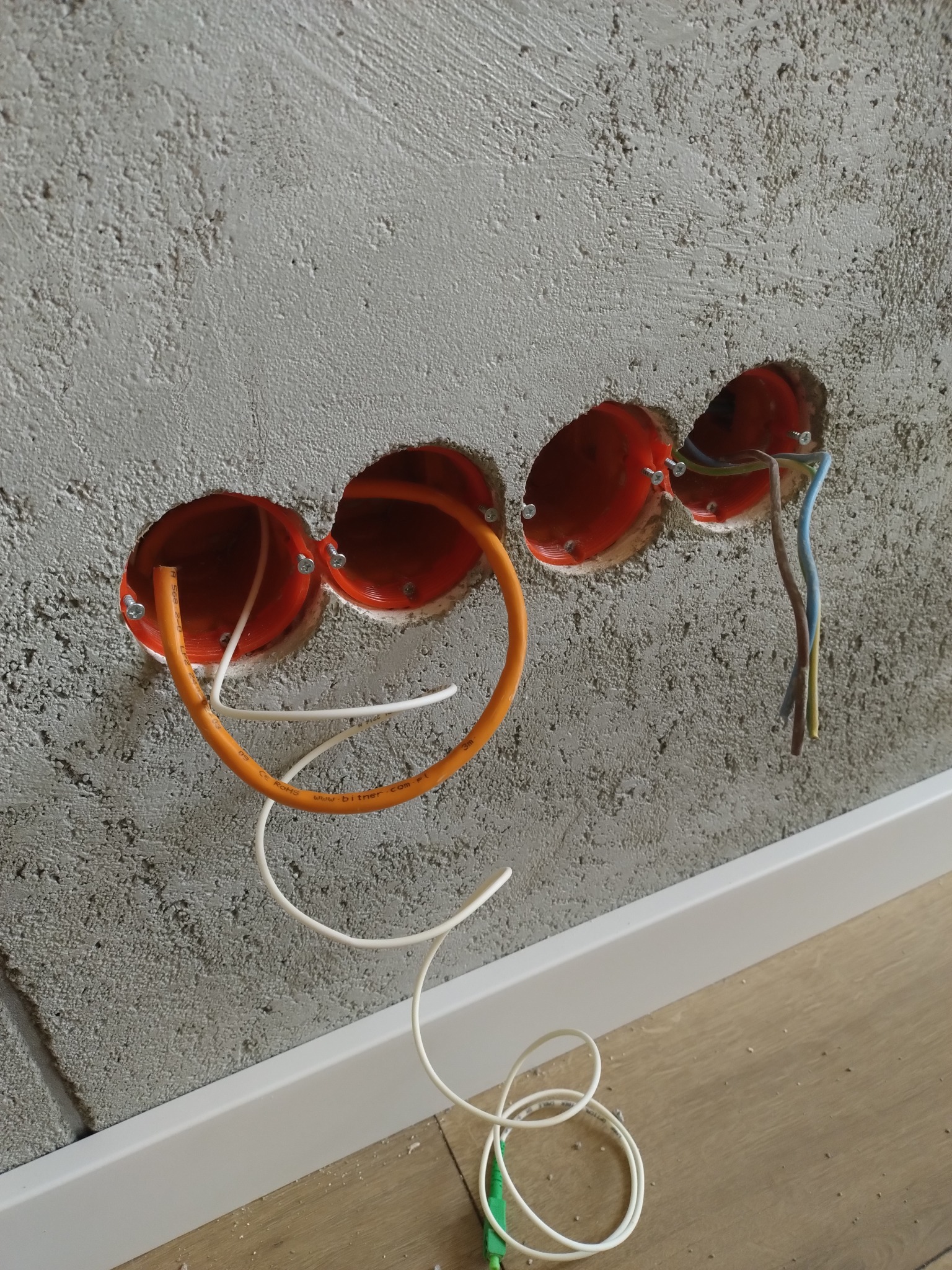 Surowa ściana z trzema okrągłymi puszkami elektrycznymi, z których wystają kable w kolorach pomarańczowym, białym i niebieskim. Widoczna listwa przypodłogowa i fragment podłogi.