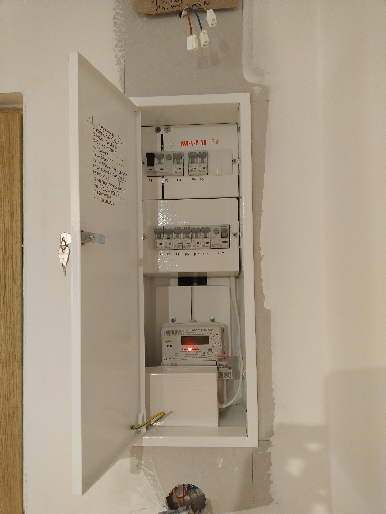 Otwarta skrzynka elektryczna z bezpiecznikami, licznikiem i listwą zaciskową na ścianie w trakcie montażu. Widoczne przewody i klucz w zamku.