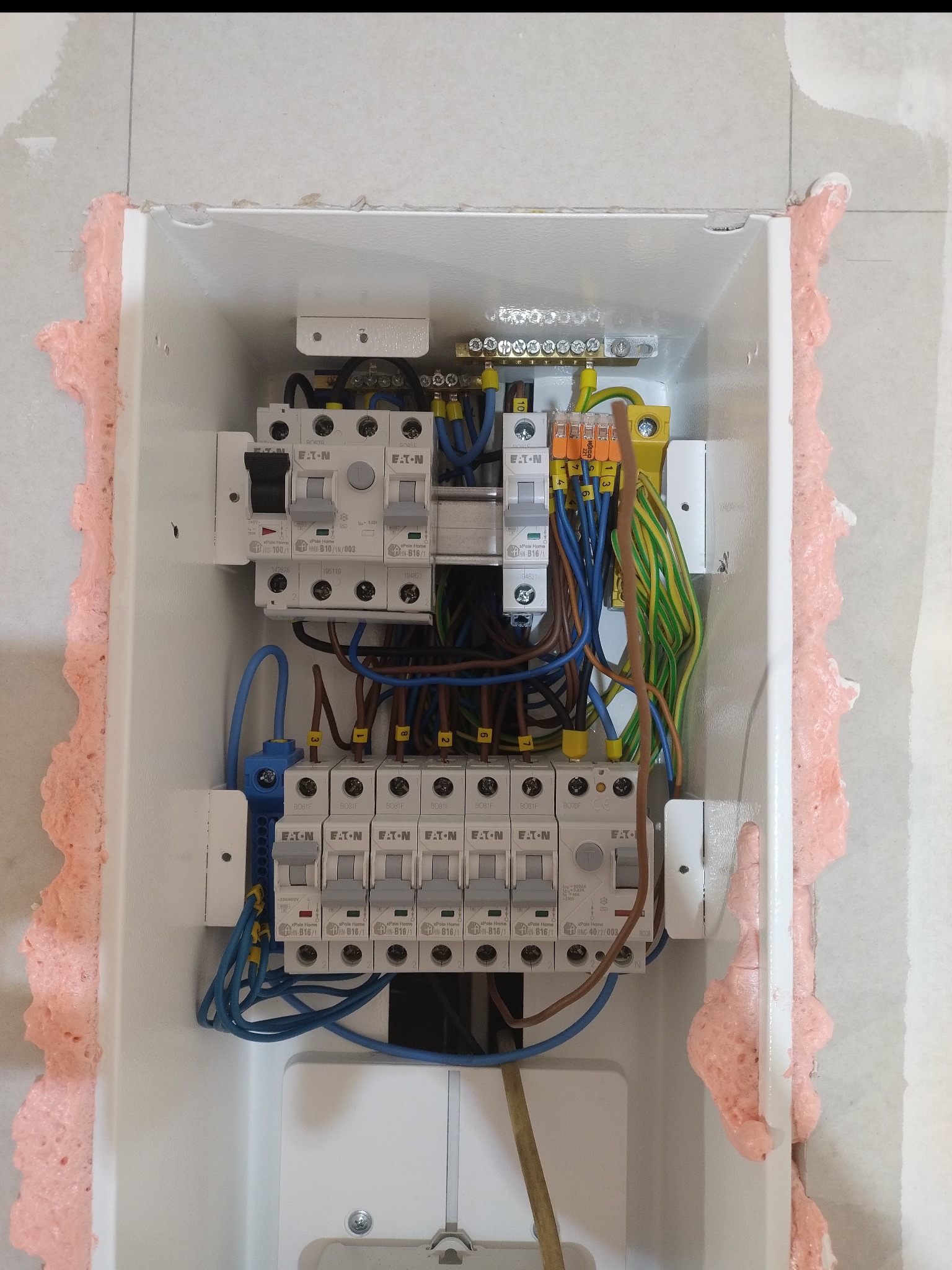 Rozdzielnica elektryczna Eaton w białej obudowie, zabezpieczona pianką montażową, z widocznymi bezpiecznikami i okablowaniem w kolorach niebieskim, brązowym i zielono-żółtym.