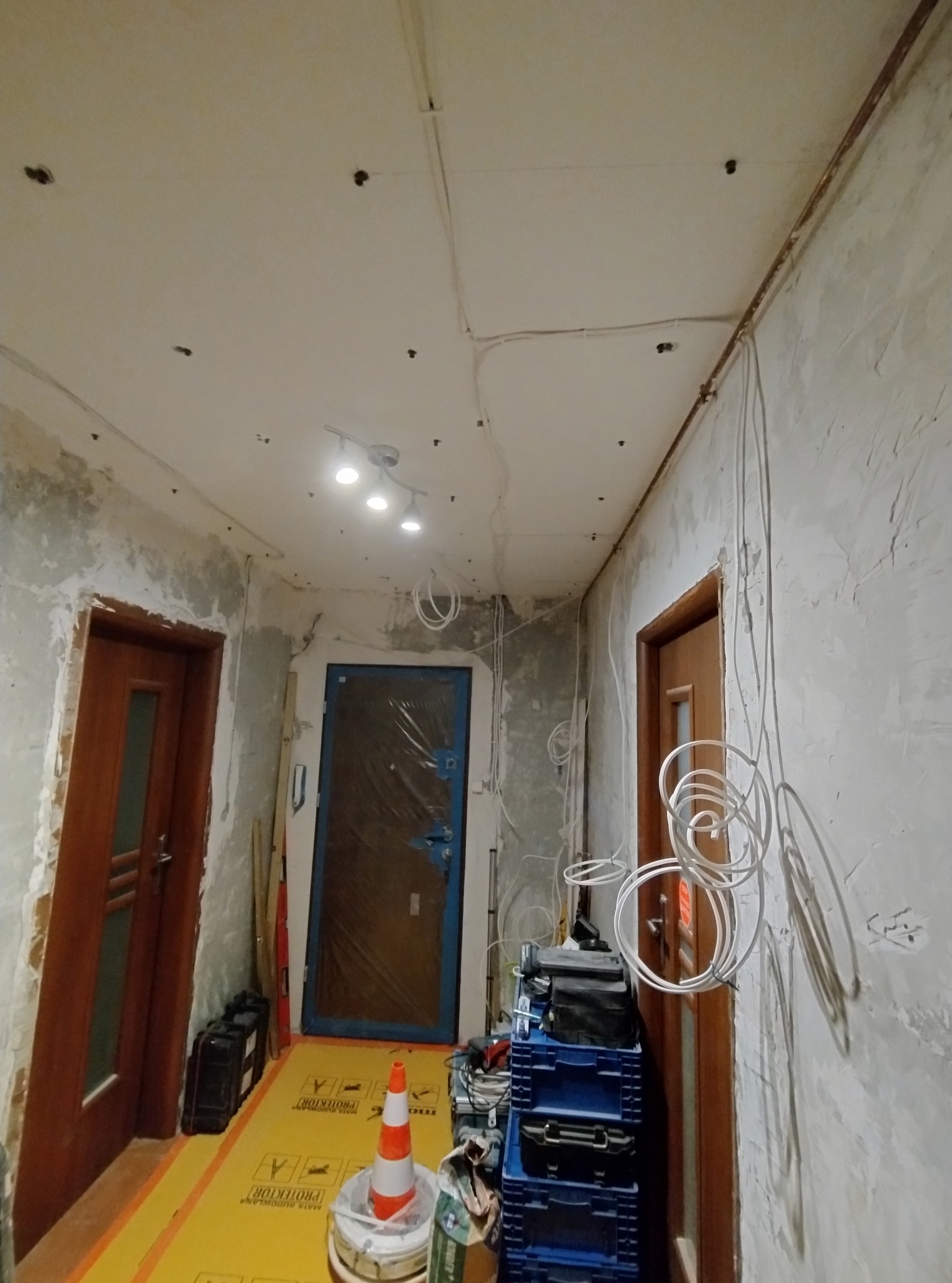 Korytarz w trakcie remontu: widoczne przewody elektryczne na suficie, ściany w stanie surowym, zabezpieczona podłoga i narzędzia. Etap prac instalacyjnych.