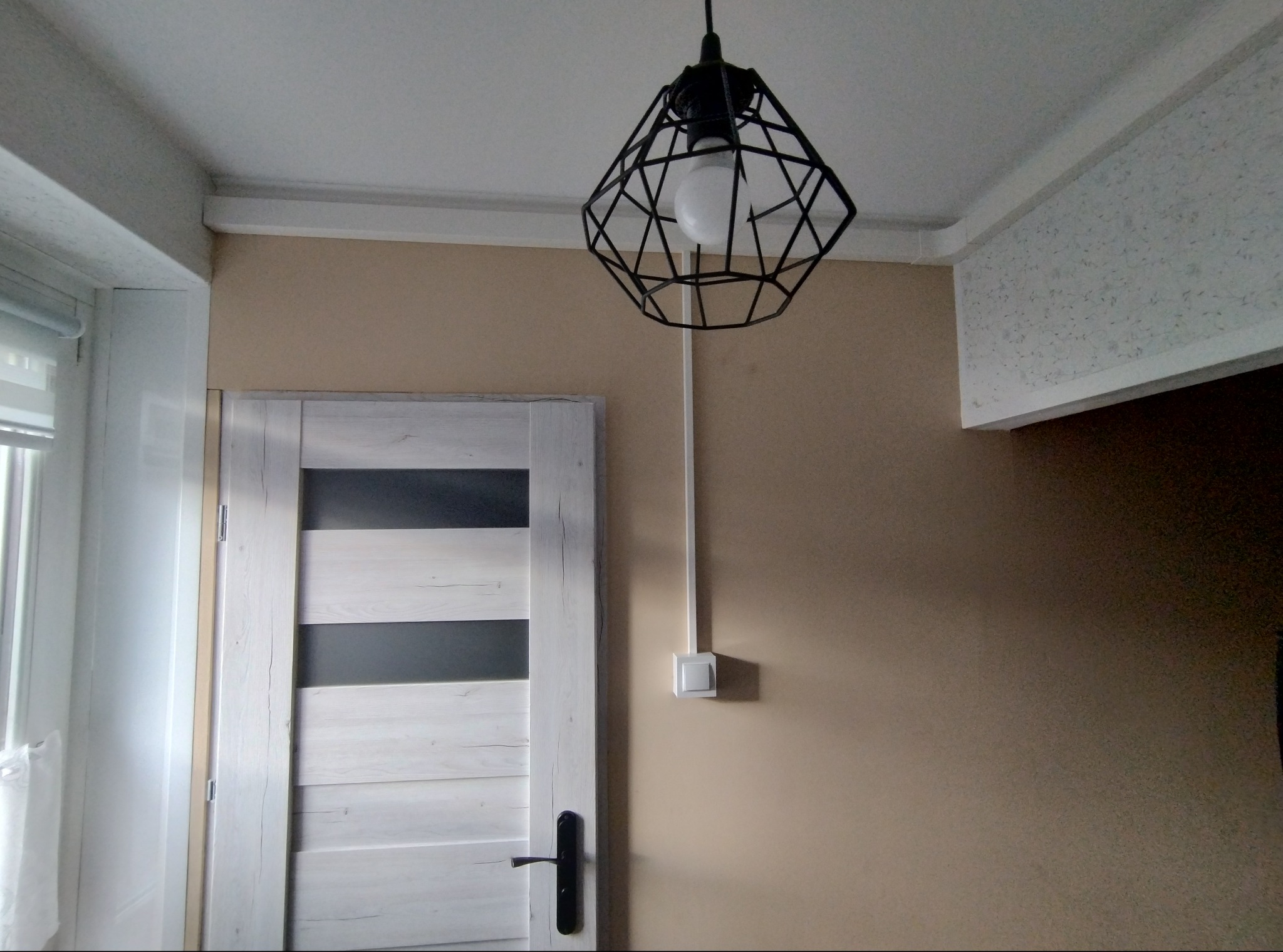 Wnętrze: białe drzwi z czarnymi wstawkami, geometryczna lampa wisząca z żarówką, beżowa ściana z włącznikiem. Widoczne fragmenty sufitu i okna.