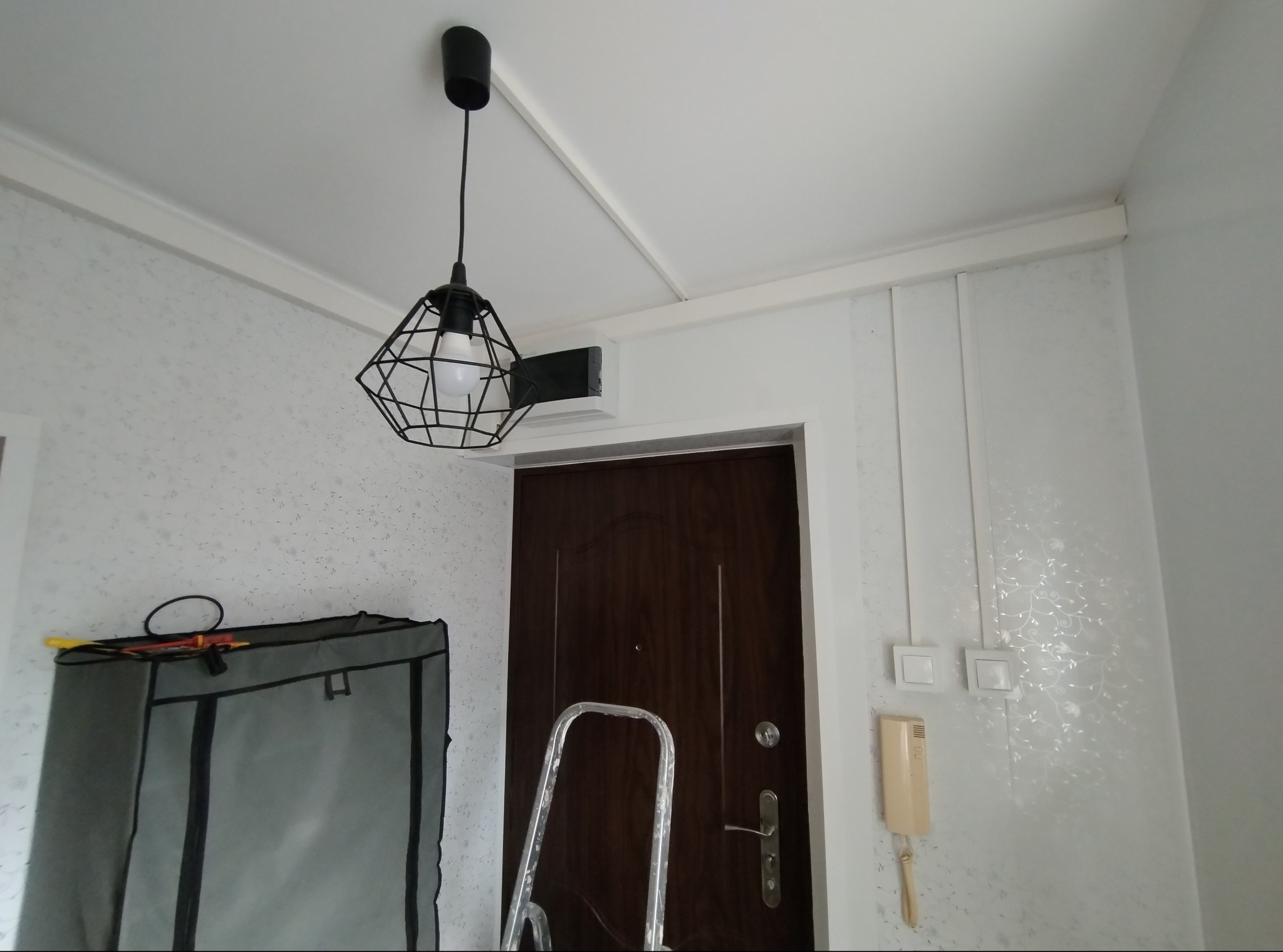 Wnętrze z zamontowaną, nowoczesną lampą wiszącą z czarnym, geometrycznym kloszem. Widoczne elementy instalacji elektrycznej i drabina malarska.