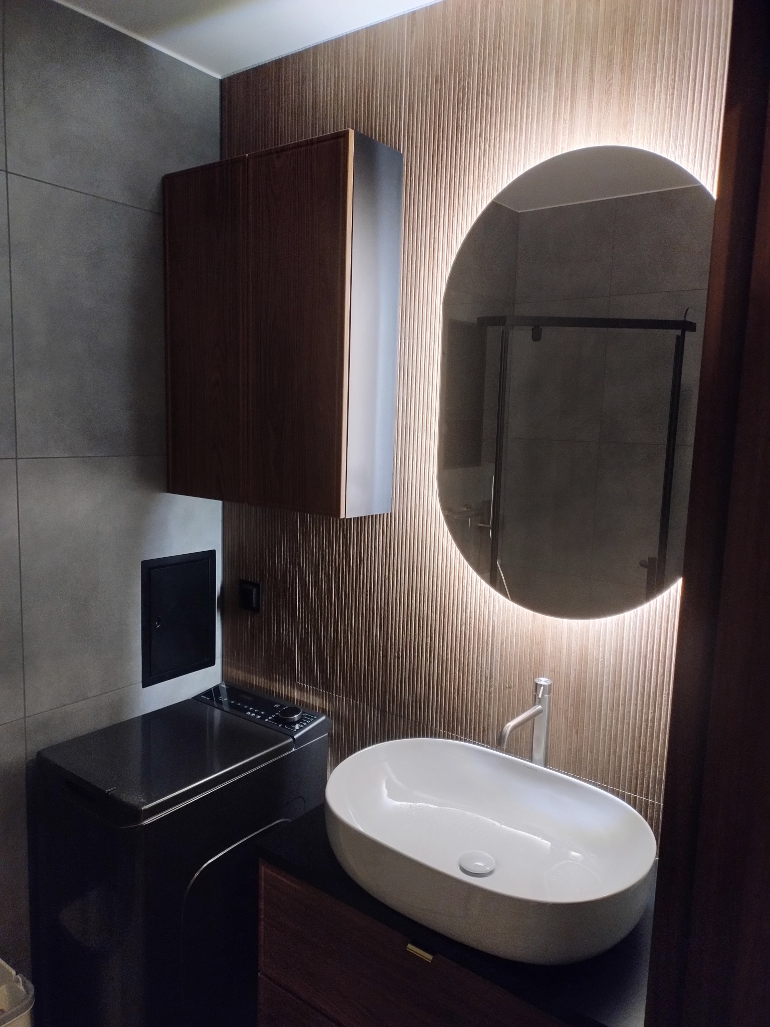 Nowoczesna łazienka z owalnym lustrem podświetlonym LED, umywalką nablatową i szafką z ciemnego drewna. Na ścianie szare płytki i dekoracyjne panele ścienne.