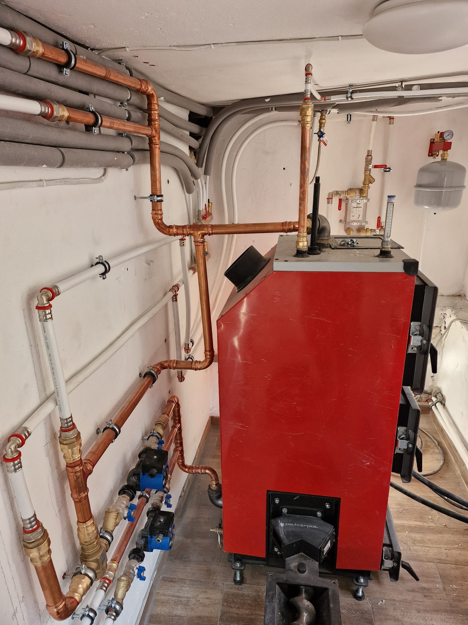 Czerwony piec grzewczy marki Padcerpol z podłączonymi rurami miedzianymi i białymi, widoczne zawory, manometry i elementy instalacji CO w kotłowni.