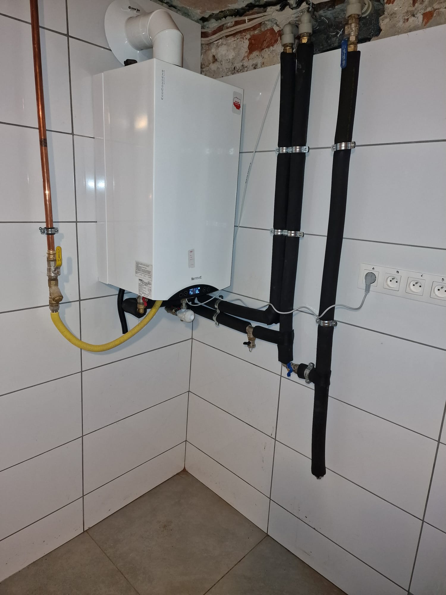 Kocioł gazowy kondensacyjny Termet zamontowany na ścianie z białych płytek, z widocznymi rurami w izolacji termicznej, przyłączem gazowym z żółtym wężem oraz gniazdkami elektrycznymi.