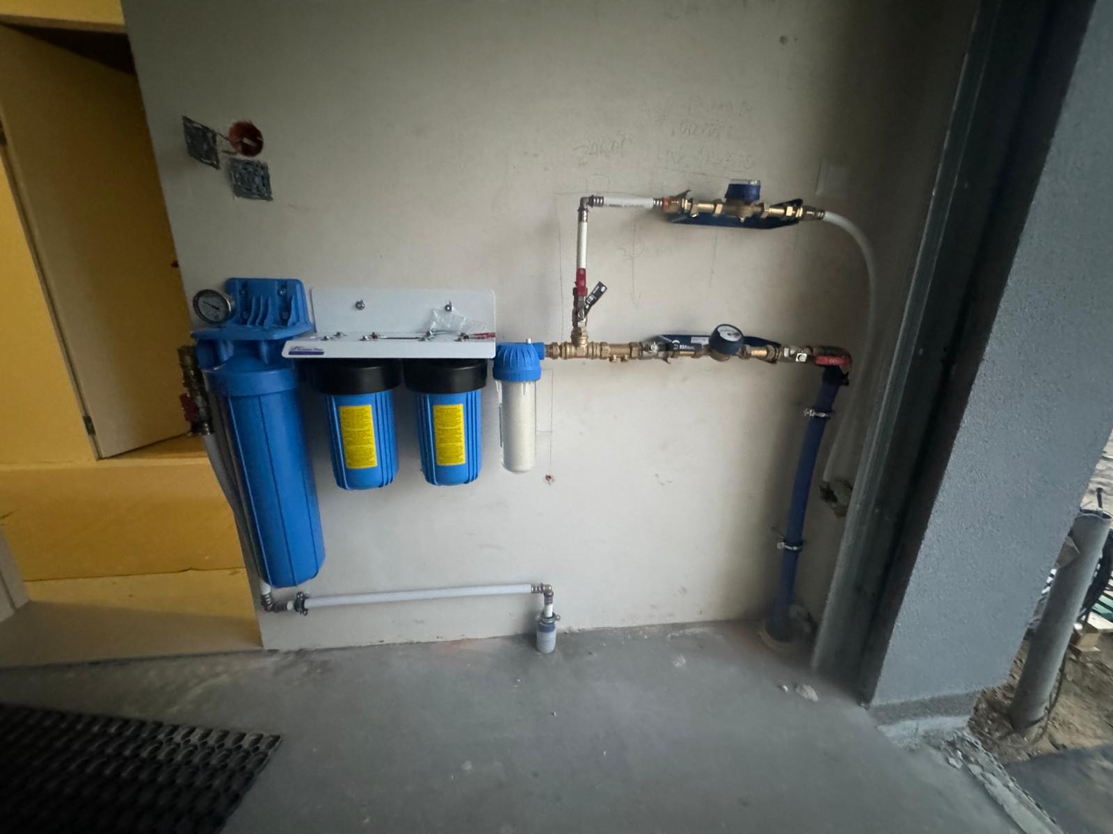 Kompleksowa instalacja filtrów wody z manometrami i zaworami, zamontowana na ścianie w pomieszczeniu gospodarczym. Widoczne rury i złączki.