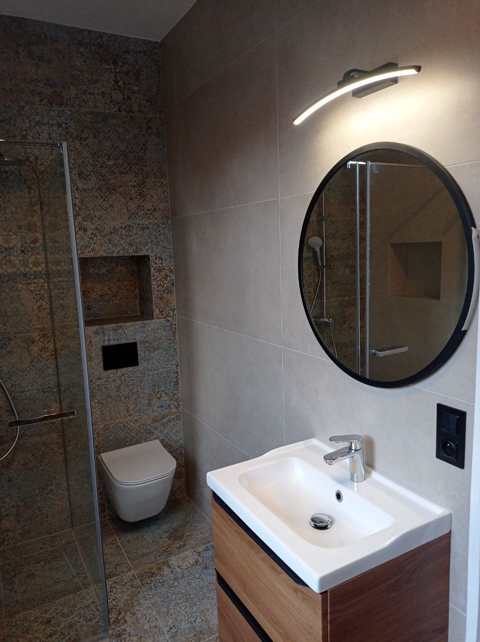 Nowoczesna łazienka z kabiną prysznicową, okrągłym lustrem z podświetleniem LED, umywalką nablatową na drewnianej szafce i toaletą podwieszaną z ukrytym spłukiwaniem.