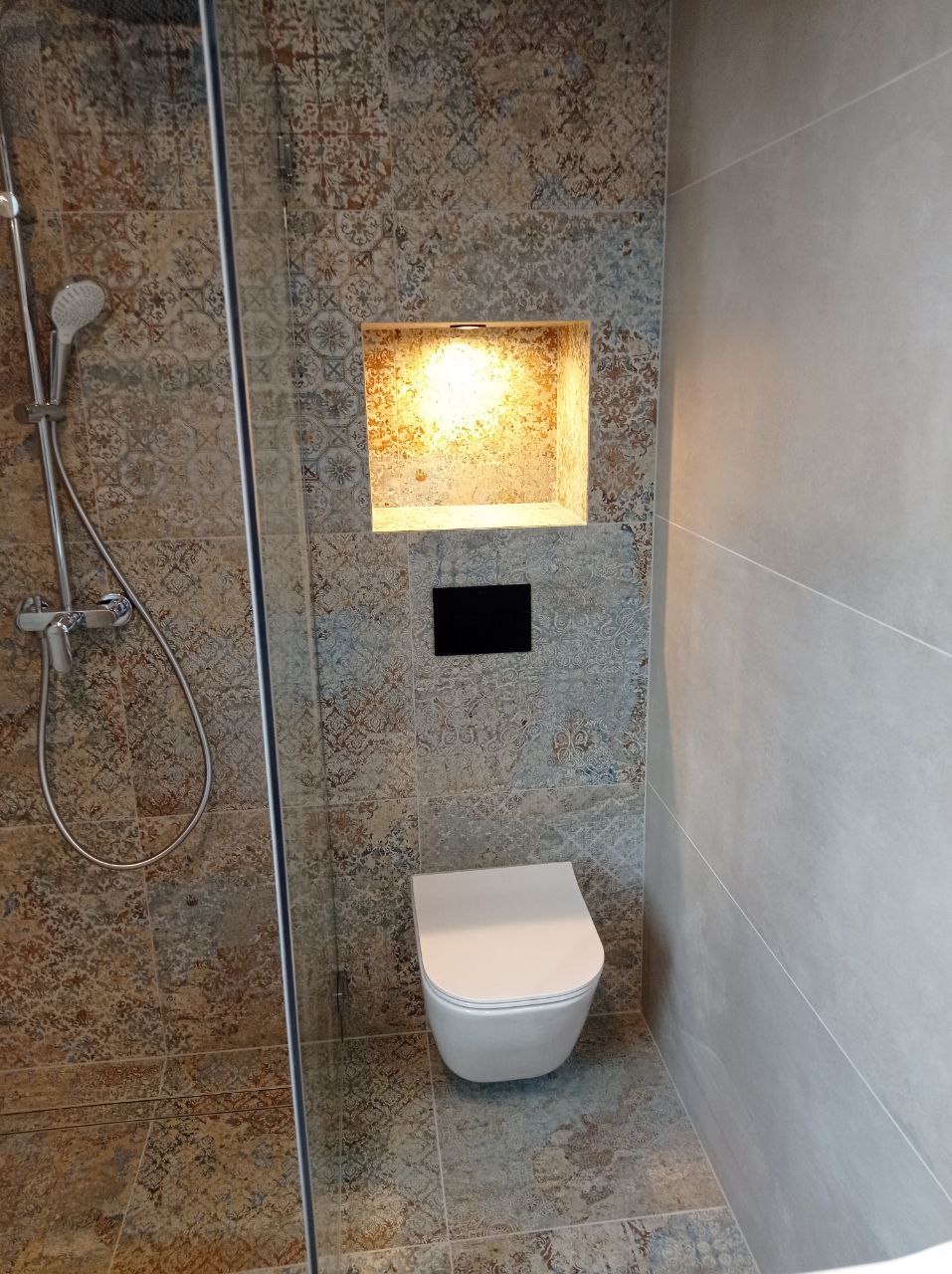 Nowoczesna łazienka z kabiną prysznicową, wiszącą toaletą i dekoracyjną wnęką z oświetleniem, wykończona płytkami w stylu patchwork i gładkimi szarymi panelami.