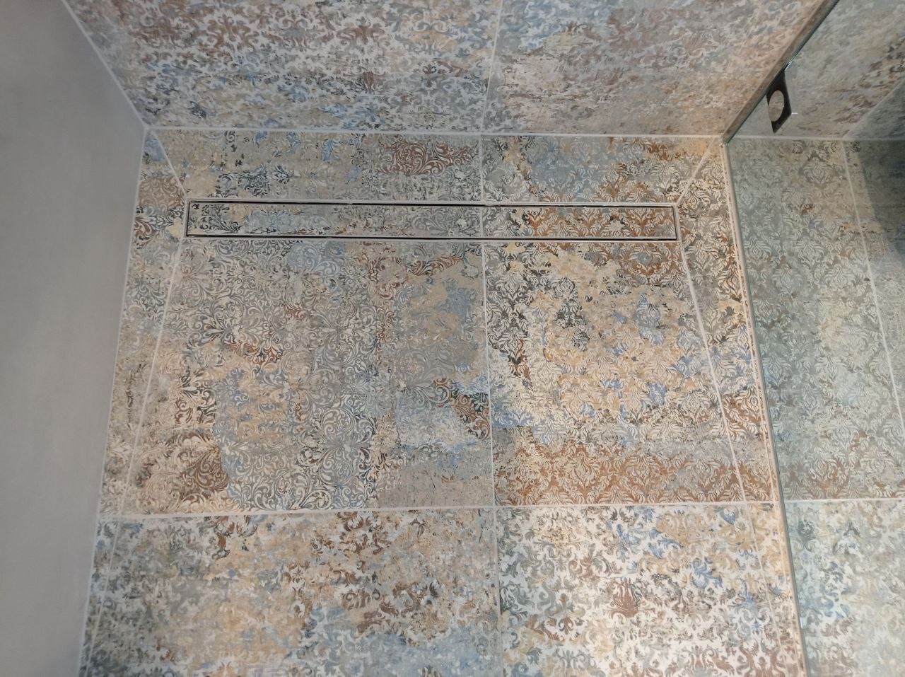 Ułożone płytki podłogowe i ścienne z dekoracyjnym wzorem w odcieniach beżu, błękitu i szarości, z odpływem liniowym w kabinie prysznicowej.