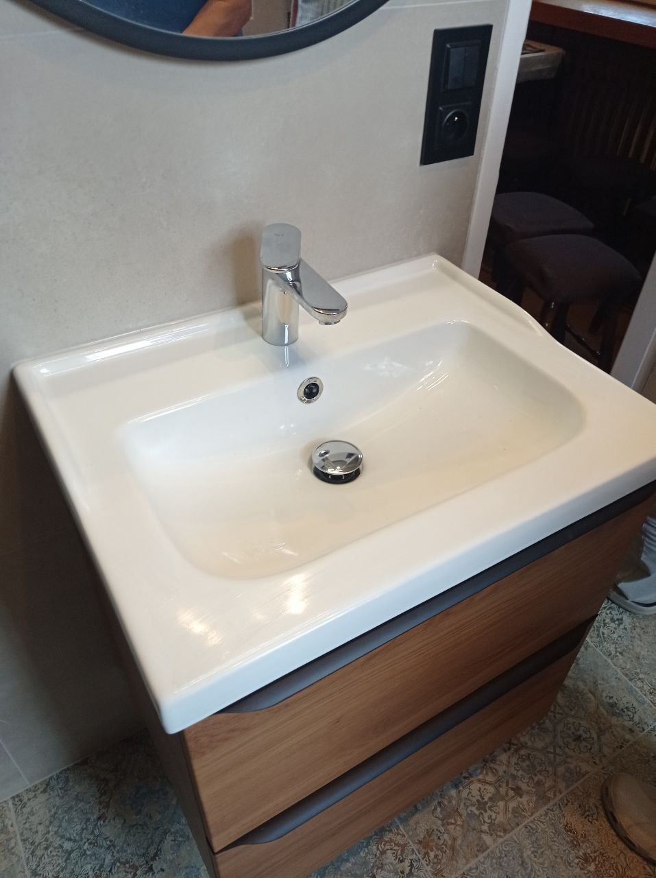 Biała umywalka z drewnianą szafką z dwoma szufladami i chromowaną baterią, widoczna część lustra i włącznik światła na ścianie.