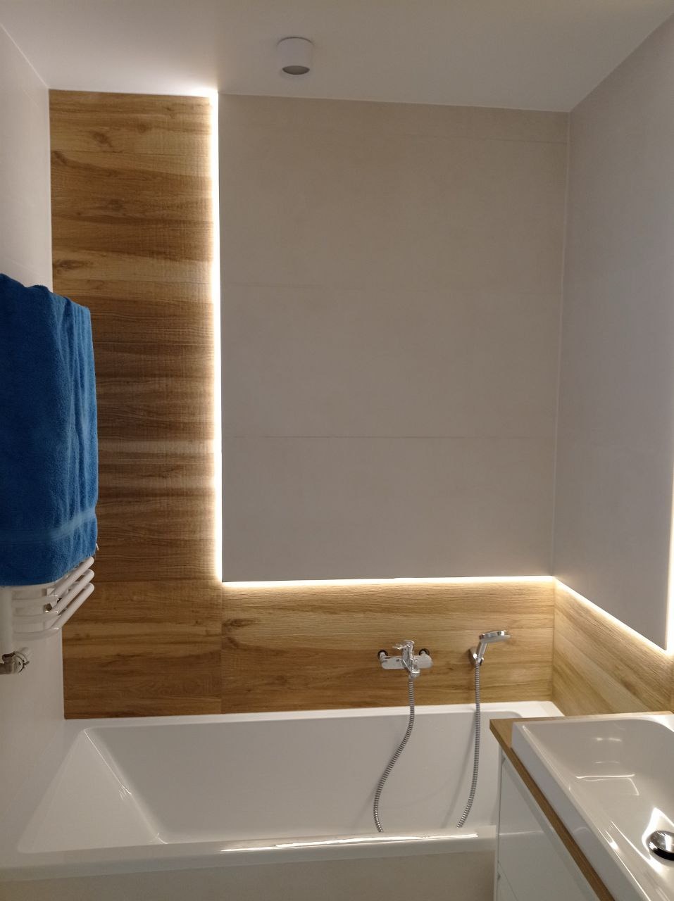 Nowoczesna łazienka z drewnopodobnymi płytkami, podświetleniem LED, białą wanną i niebieskim ręcznikiem na grzejniku.