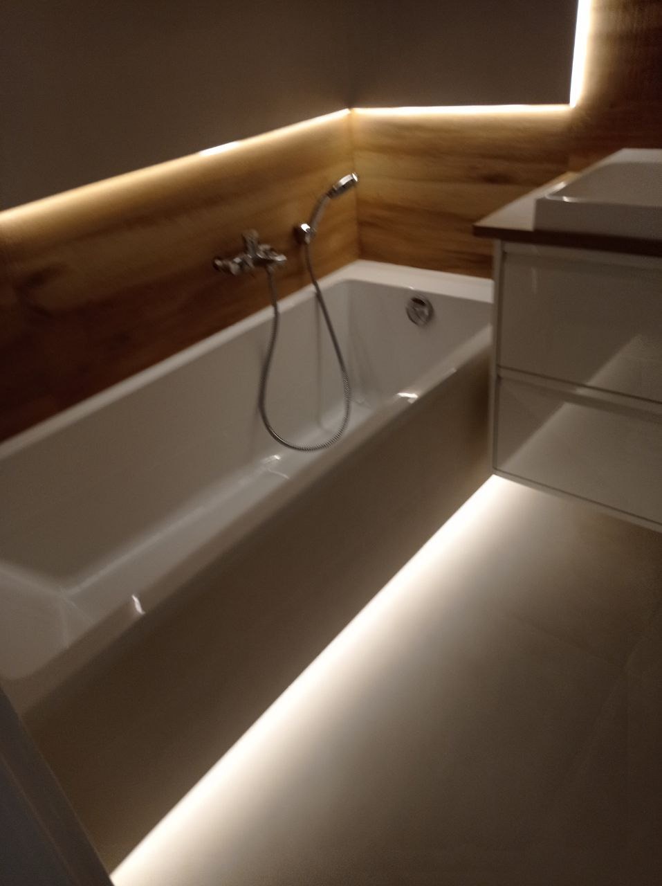 Biała wanna z chromowaną baterią i prysznicem, oświetlenie LED pod szafką umywalkową i nad drewnopodobną okładziną ścienną, minimalistyczny design łazienki.