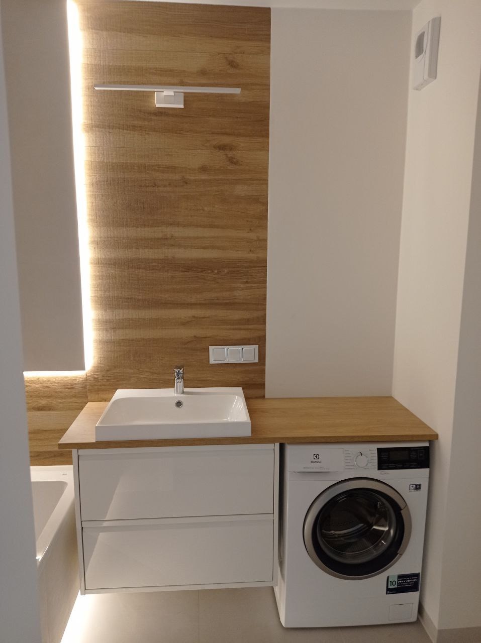 Nowoczesna łazienka z drewnopodobną okładziną ścienną, białą umywalką nablatową z szafką i pralką Electrolux z blatem w kolorze drewna.