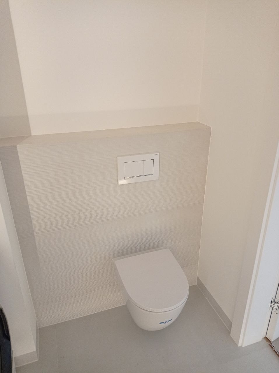 Nowoczesna toaleta podwieszana z białym panelem sterującym na ścianie z delikatną fakturą, szare płytki podłogowe i minimalistyczny design.