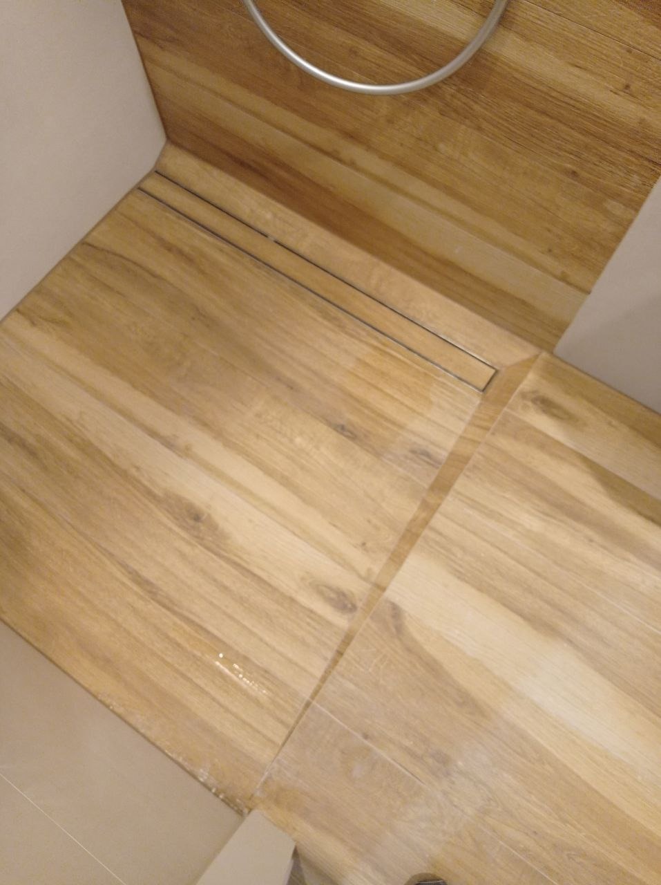 Drewnopodobne płytki podłogowe w łazience z odpływem liniowym, widoczne fugi i fragment kabiny prysznicowej.