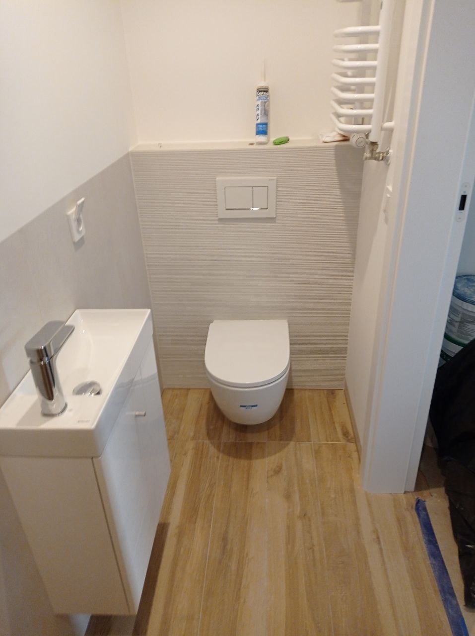 Nowoczesna łazienka z podwieszaną toaletą, umywalką z szafką i grzejnikiem drabinkowym, wykończona płytkami imitującymi drewno na podłodze i białymi płytkami strukturalnymi na ścianach.