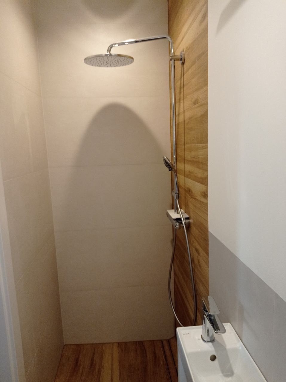 Nowoczesna kabina prysznicowa z deszczownicą i baterią, wykończona płytkami imitującymi drewno na jednej ze ścian, z minimalistyczną umywalką w rogu.