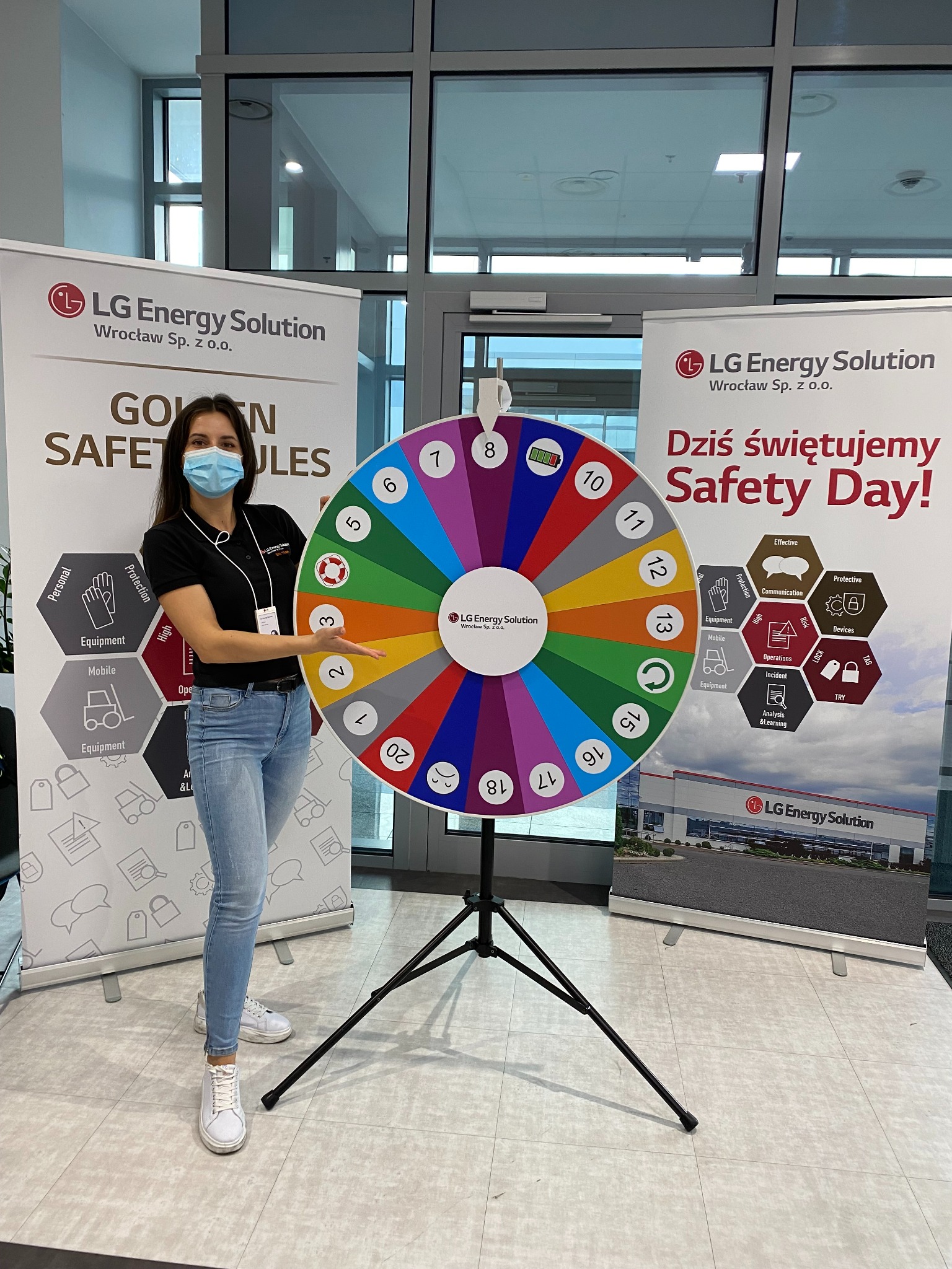 Kobieta w maseczce prezentuje koło fortuny z numerami, obok roll-upy LG Energy Solution z hasłami BHP. Dzień Bezpieczeństwa w firmie.