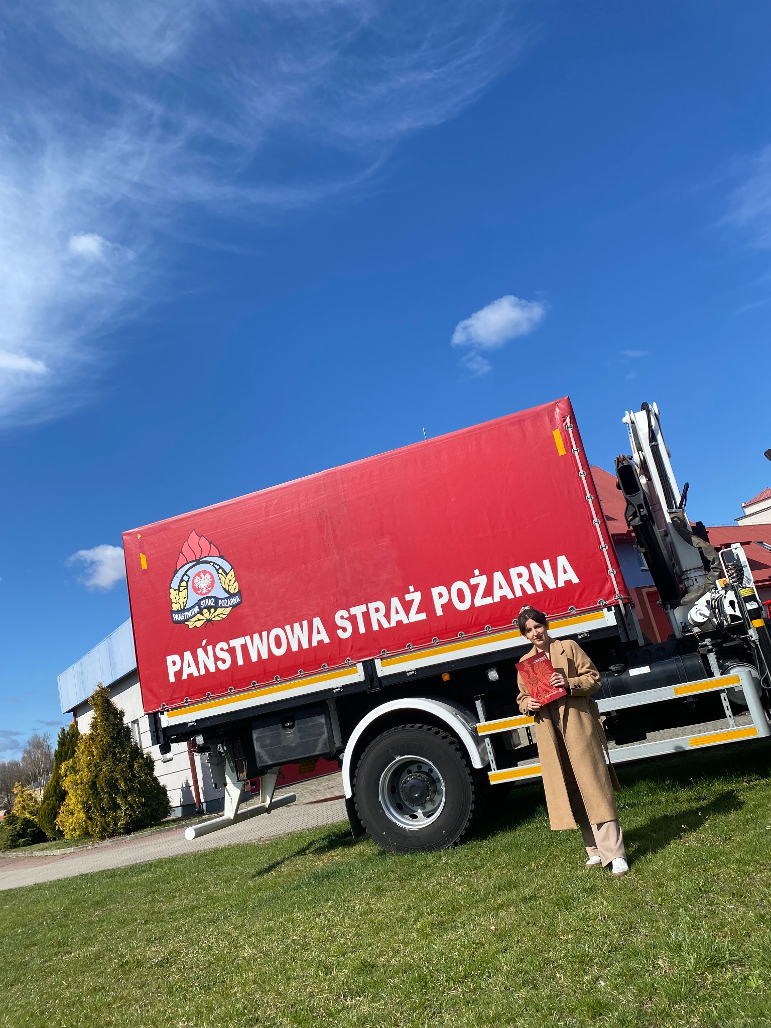 Kobieta w płaszczu prezentuje materiały szkoleniowe na tle czerwonej ciężarówki Państwowej Straży Pożarnej w słoneczny dzień. Błękitne niebo z chmurami w tle.
