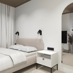 INTERIOR DESIGN Julia Jarząbkiewicz - Jasna sypialnia z łóżkiem kontynentalnym, lampkami nocnymi na ścianie i szafką nocną z szufladą; widok przez łukowe przejście do dalszej części mieszkania.