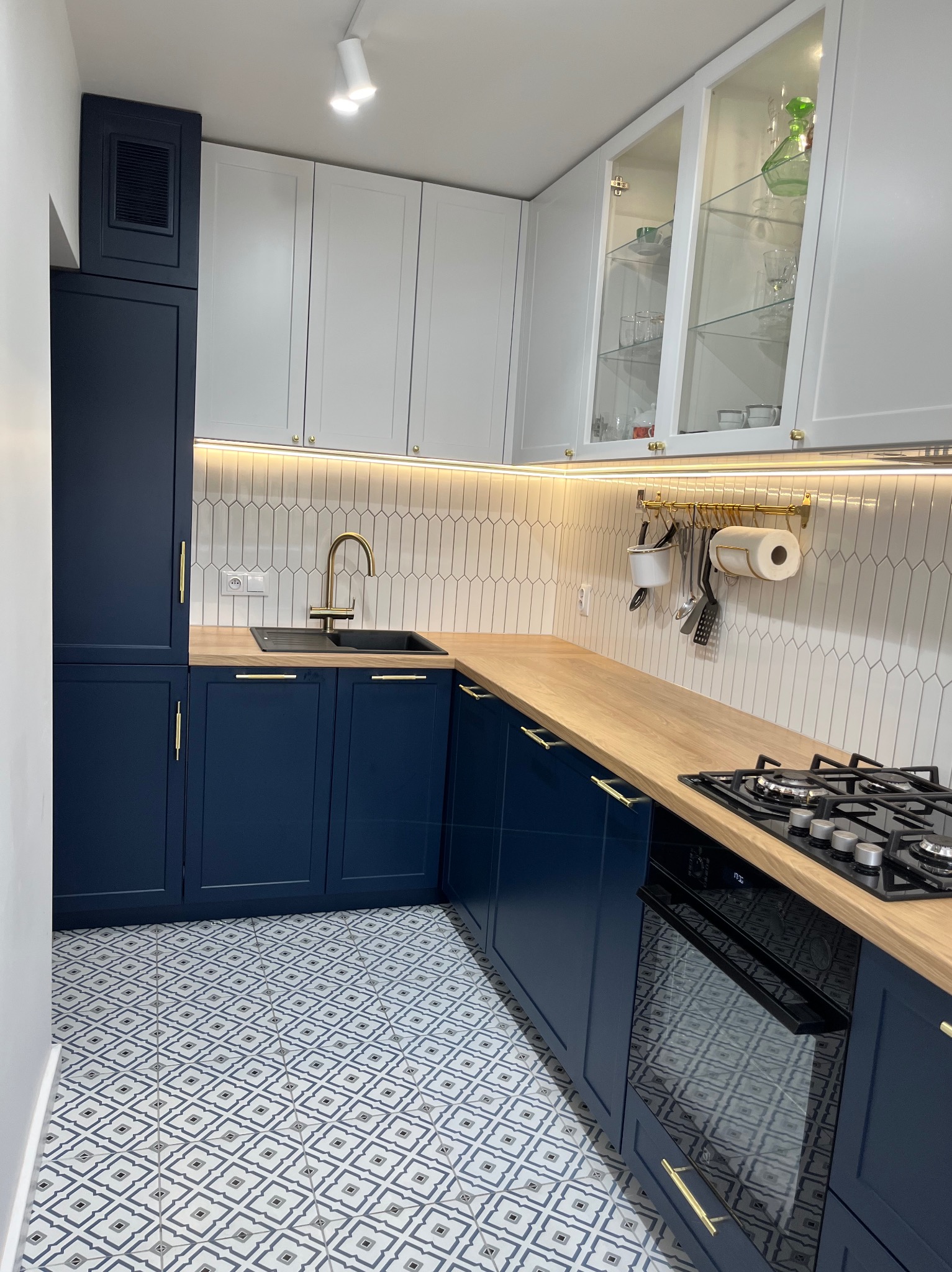 Granatowo-biała kuchnia z drewnianym blatem w Gdyni, z wzorzystą podłogą, złotymi detalami i oświetleniem LED pod szafkami.