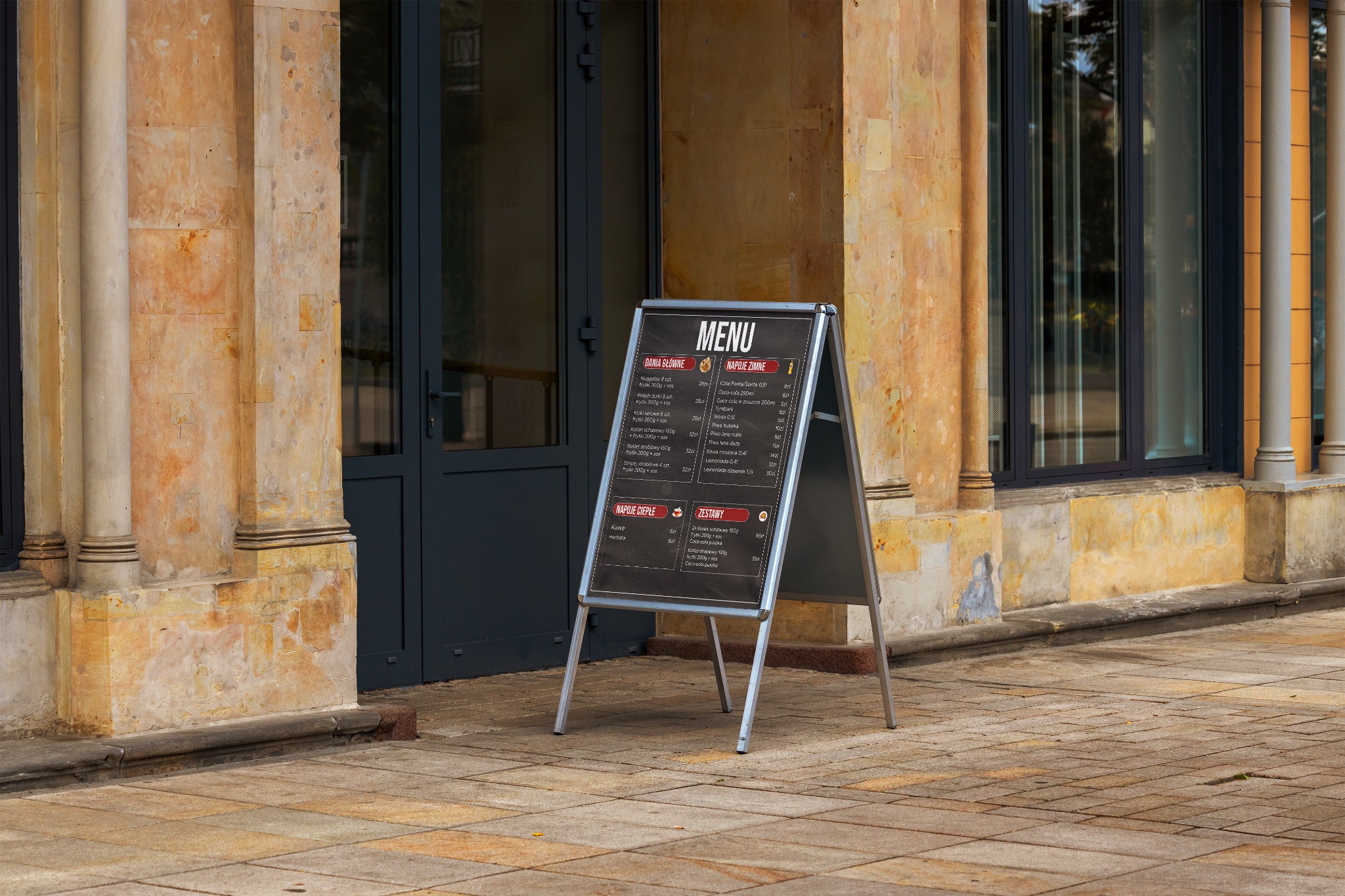 Dwustronny, srebrny stojak reklamowy z menu restauracji w Woli Łaszewskiej, ustawiony na chodniku przed wejściem do lokalu z kolumnami w tle.