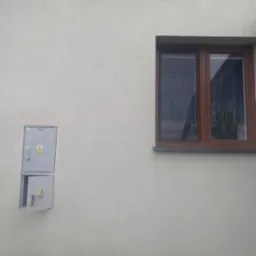 Jasna elewacja budynku z brązowym oknem i szarą szafką elektryczną, której dolne drzwiczki są otwarte. Widoczne znaki ostrzegawcze.