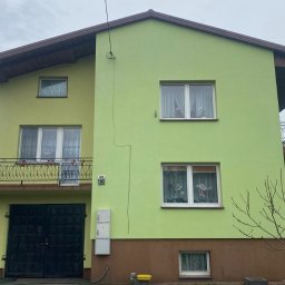 Radosław Badowski - Dwukondygnacyjny dom z zieloną elewacją i ciemnobrązowym dachem, widoczne okna z firankami i balkon z ozdobną balustradą, ciemne dwuskrzydłowe drzwi garażowe.