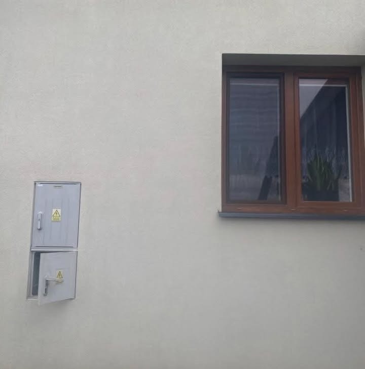 Jasna elewacja budynku z brązowym oknem i szarą szafką elektryczną, której dolne drzwiczki są otwarte. Widoczne znaki ostrzegawcze.