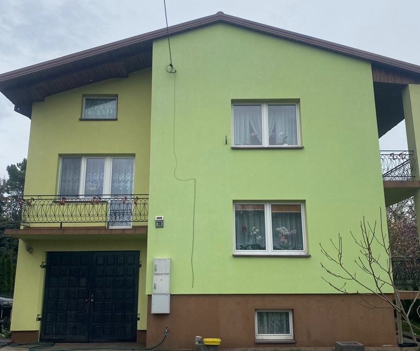 Dwukondygnacyjny dom z zieloną elewacją i ciemnobrązowym dachem, widoczne okna z firankami i balkon z ozdobną balustradą, ciemne dwuskrzydłowe drzwi garażowe.