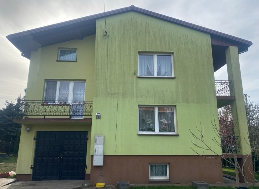 Dwukondygnacyjny dom z zieloną elewacją i brązowym cokołem, widoczne ślady zabrudzeń i zacieków na ścianach. Balkon z czarną balustradą.