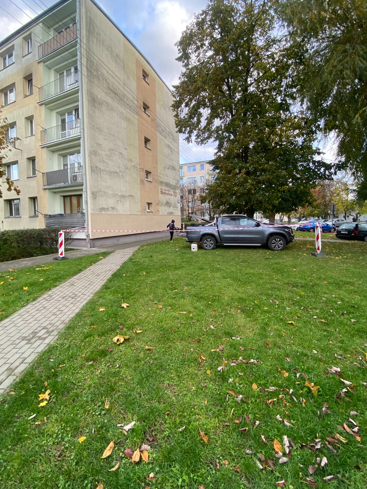 Przygotowania do prac elewacyjnych: budynek zabezpieczony taśmą, obok zaparkowany samochód z otwartą paką i widocznym wiadrem. Jesienna sceneria.