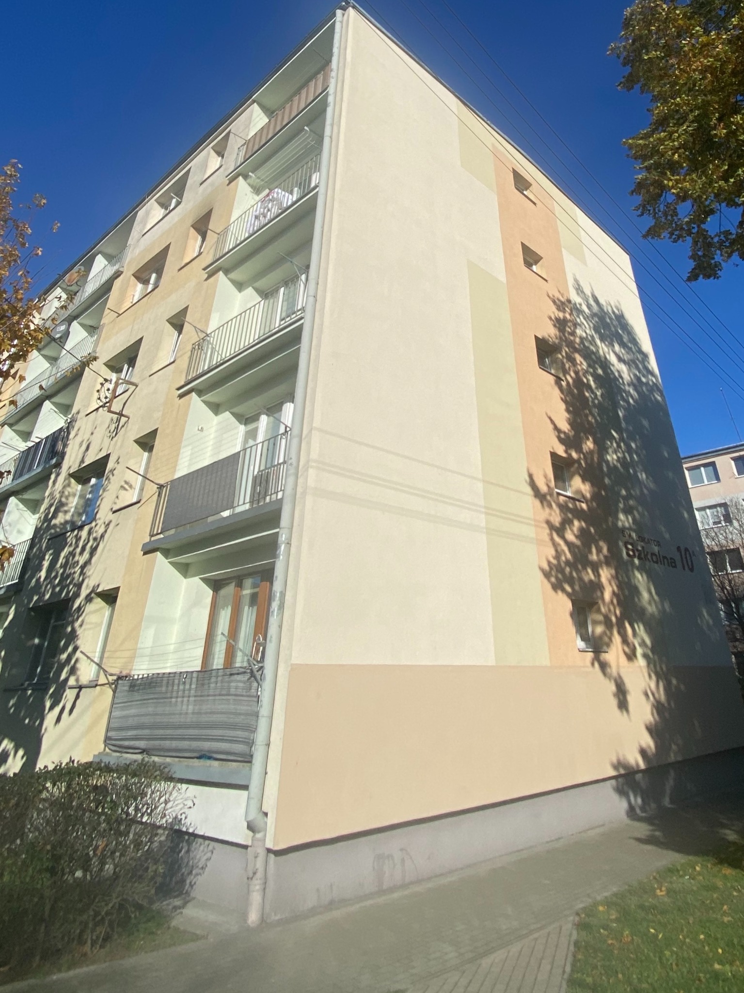 Wielopiętrowy budynek po renowacji elewacji, z widocznymi balkonami i fragmentem zieleni. Na ścianie napis 'Szkolna 10'. Ujęcie z dołu, podkreślające wysokość budynku.