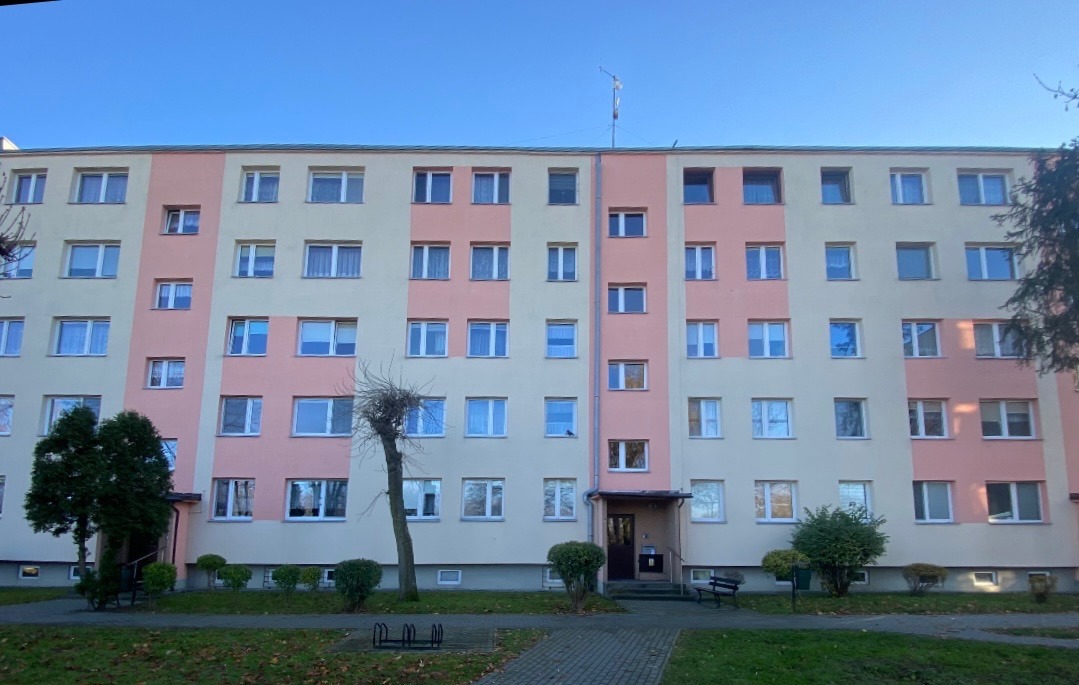 Wielopiętrowy budynek mieszkalny z elewacją w odcieniach beżu i różu, widoczny z poziomu chodnika. Na pierwszym planie zieleń i drzewa.