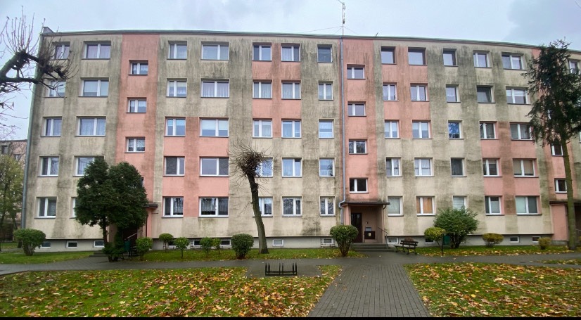 Wielopiętrowy budynek z widocznymi śladami zabrudzeń na elewacji, zieleń przed budynkiem, pochmurne niebo. Widoczne zacieki na ścianach.