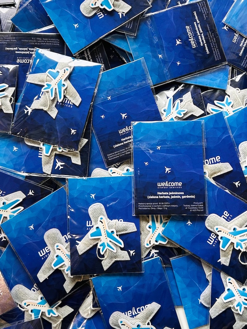 Stos niebieskich saszetek herbaty 'Welcome Airport Services' z motywem samolotu i dołączonym filcowym breloczkiem w kształcie samolotu.