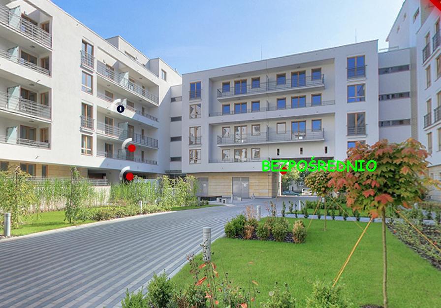 Nowoczesny apartamentowiec z balkonami i zadbanym terenem zielonym z chodnikiem z kostki brukowej. Na elewacji widoczny napis 'BEZPOŚREDNIO'.