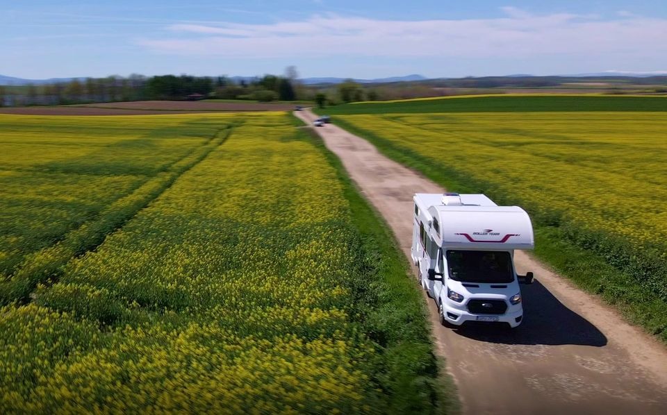 Biały kamper marki Roller Team na polnej drodze, otoczony kwitnącym na żółto polem rzepaku, z widocznymi w tle wzgórzami pod błękitnym niebem.