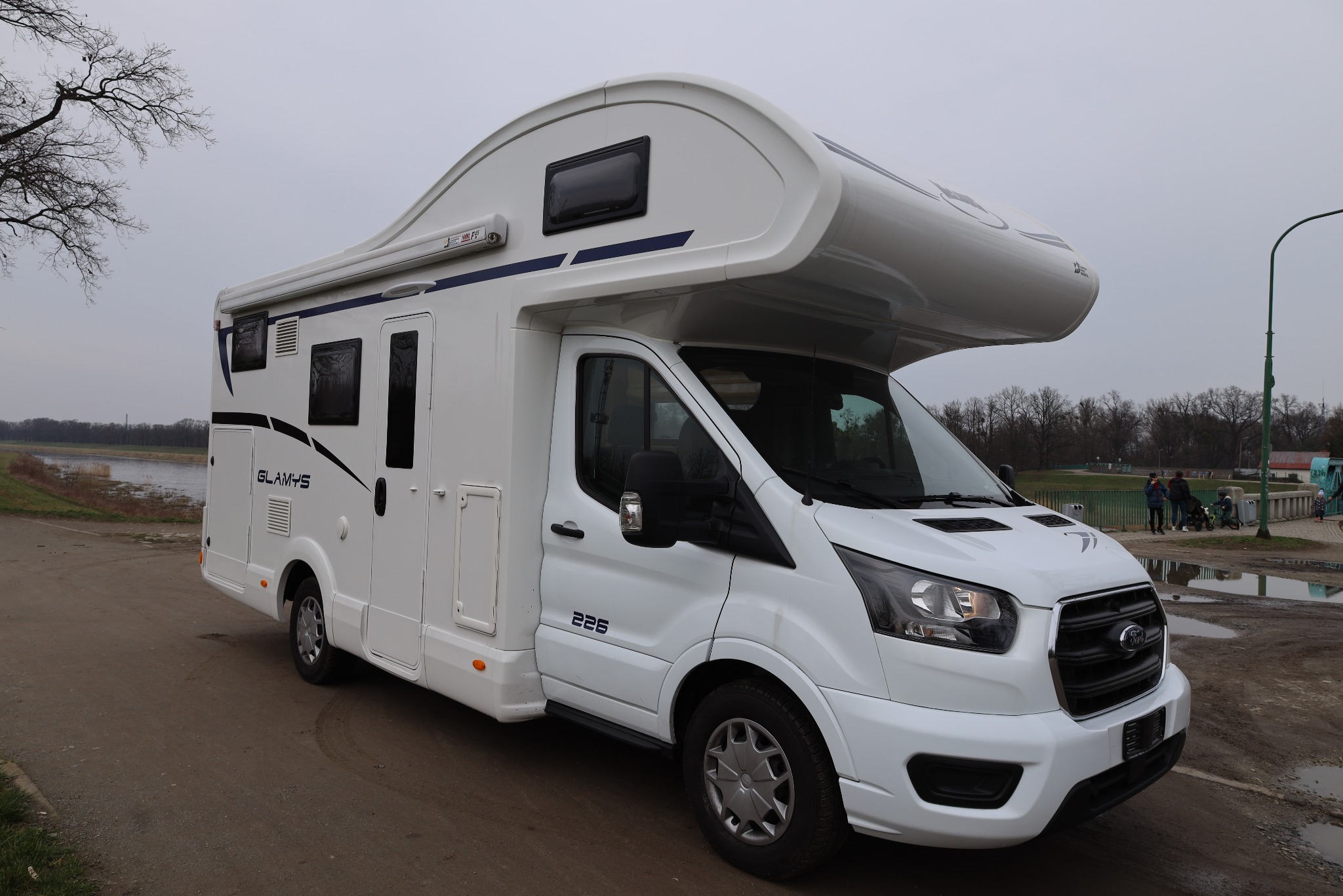 Biały kamper McLouis Glamys 226 na szutrowym parkingu, z rzeką i drzewami w tle, uchwycony w pochmurny dzień.