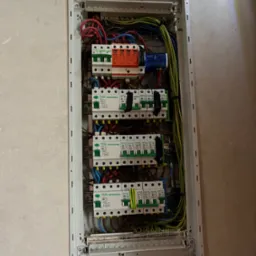 Szczegółowe ujęcie rozdzielnicy elektrycznej w jasnej obudowie, z widocznymi bezpiecznikami, przewodami w różnych kolorach i elementami zabezpieczeń.