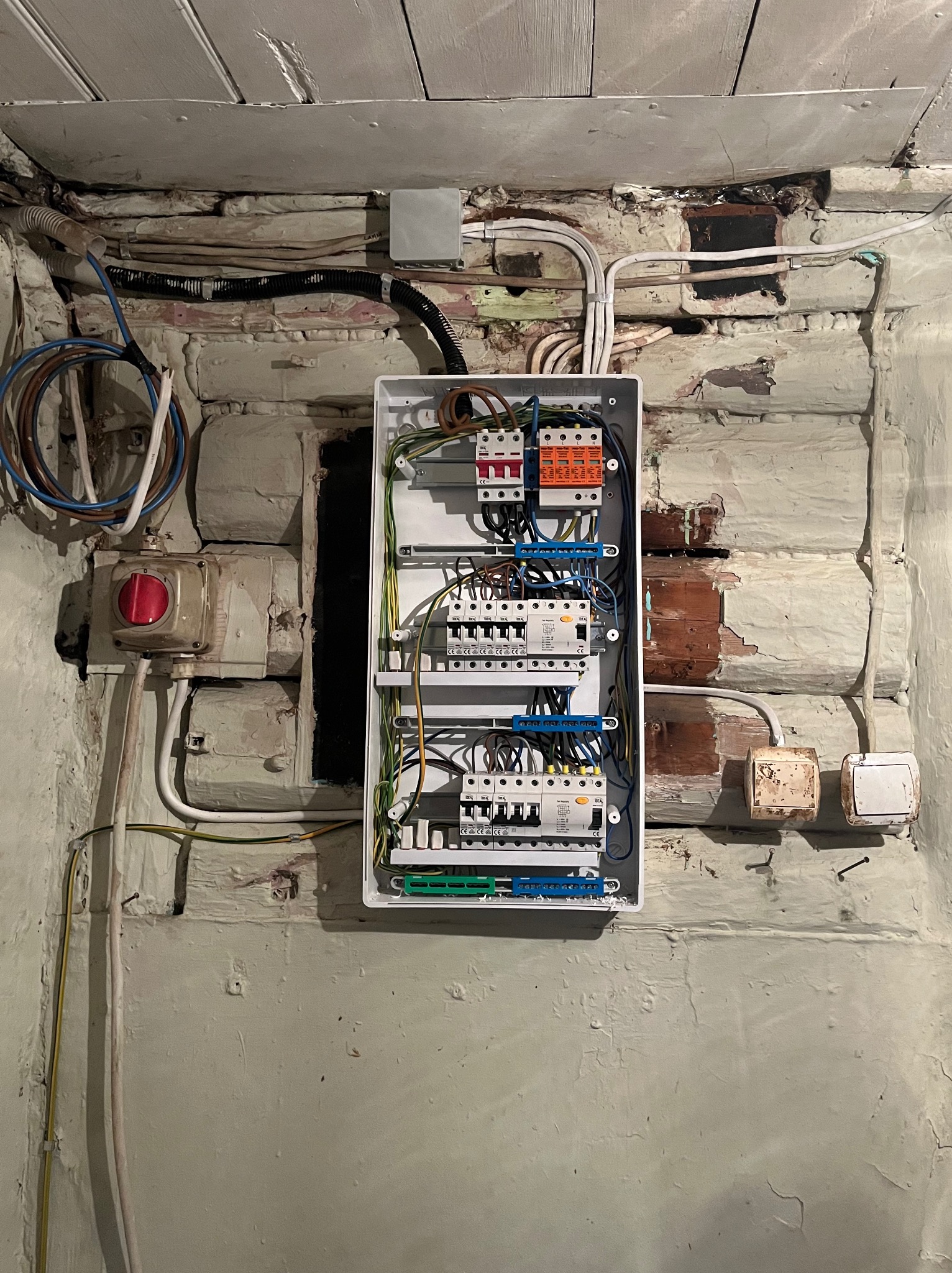 Rozdzielnica elektryczna w białej obudowie zamontowana na starej, pomalowanej ceglanej ścianie. Widoczne kable i przewody elektryczne oraz wyłączniki.