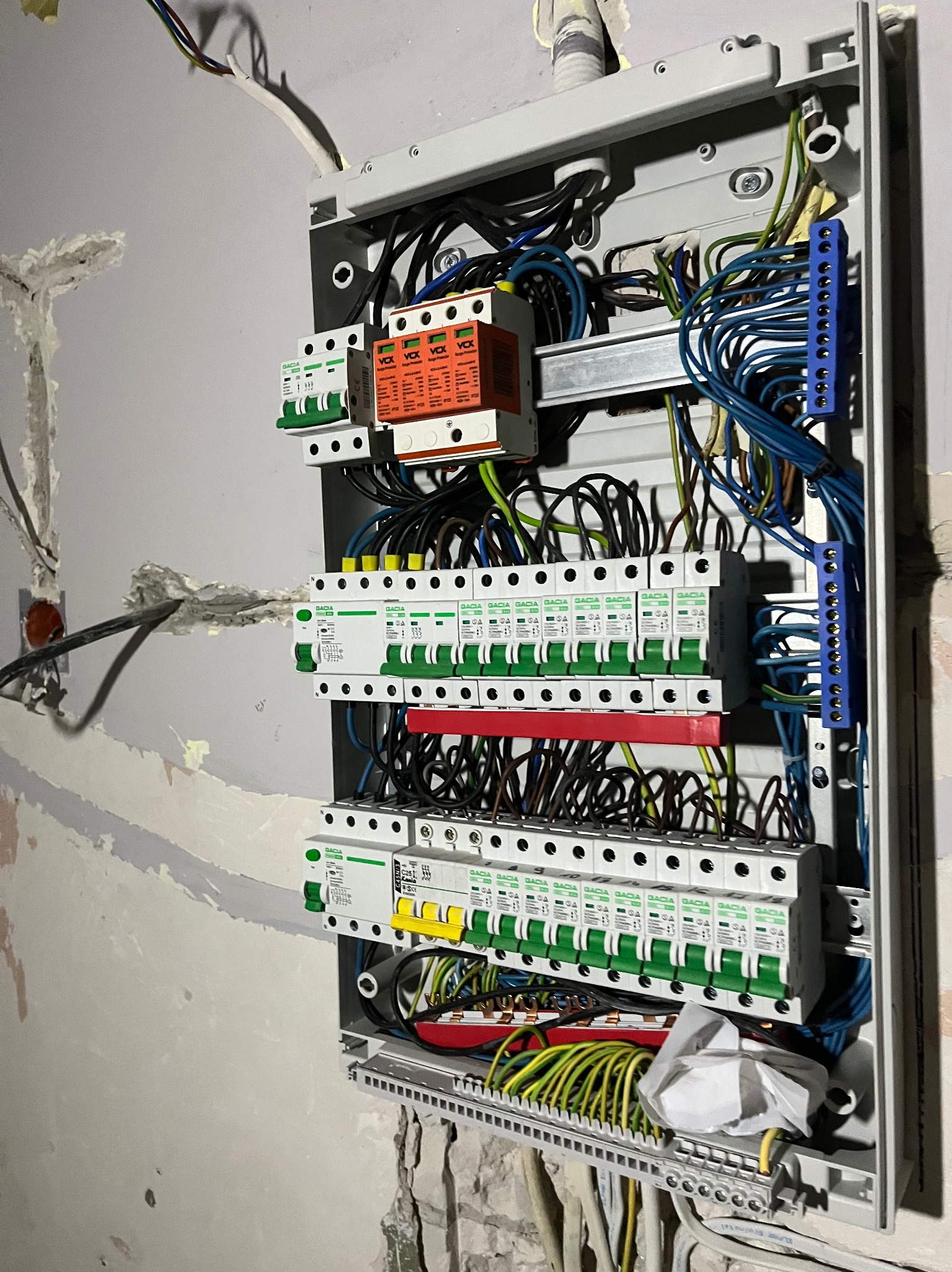 Otwarta skrzynka rozdzielcza z zainstalowanymi bezpiecznikami i okablowaniem na surowej ścianie. Widoczne kable w różnych kolorach oraz elementy montażowe wewnątrz obudowy.